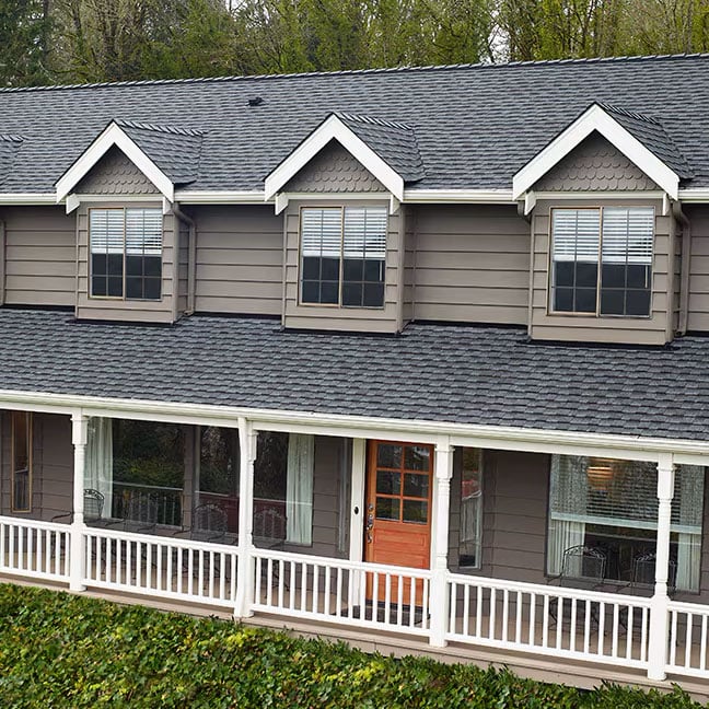 Two-story home with GAF Timberline UHDZ Pewter Gray architectural shingles, multiple dormer windows, a wraparound porch with white railings, and a vibrant orange front door, surrounded by greenery