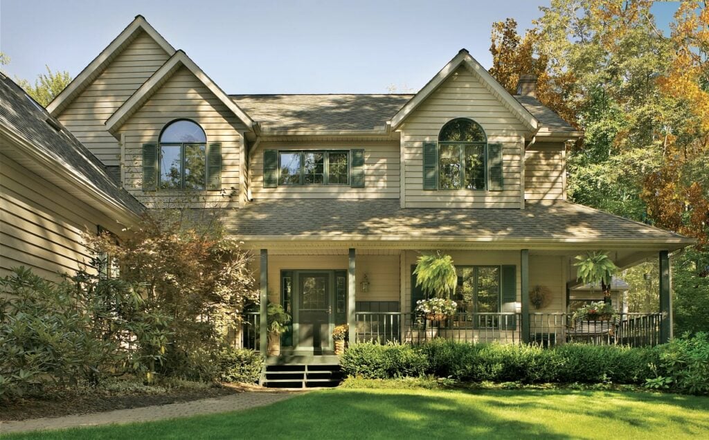 Beautiful two-story home featuring Prodigy® insulated vinyl siding in a warm beige tone, complemented by green shutters and a welcoming front porch with lush landscaping.